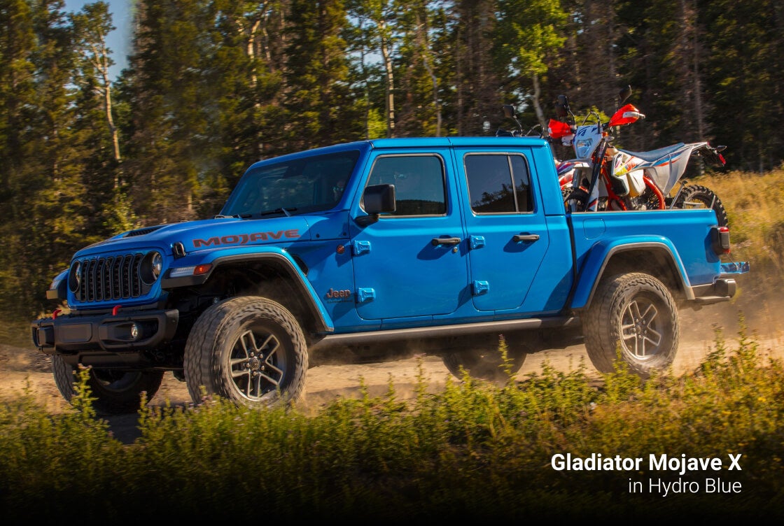2026 Jeep Gladiator Mojave X in Hydro Blue hauling dirt bikes on a forest trail.